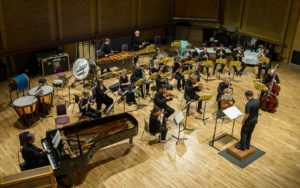 The Birmingham Contemporary Music Group play in an orchestral formation in a hall with hard wood floors. There are various instruments in the group, piano, stirings, brass, horns and woodwind. 
