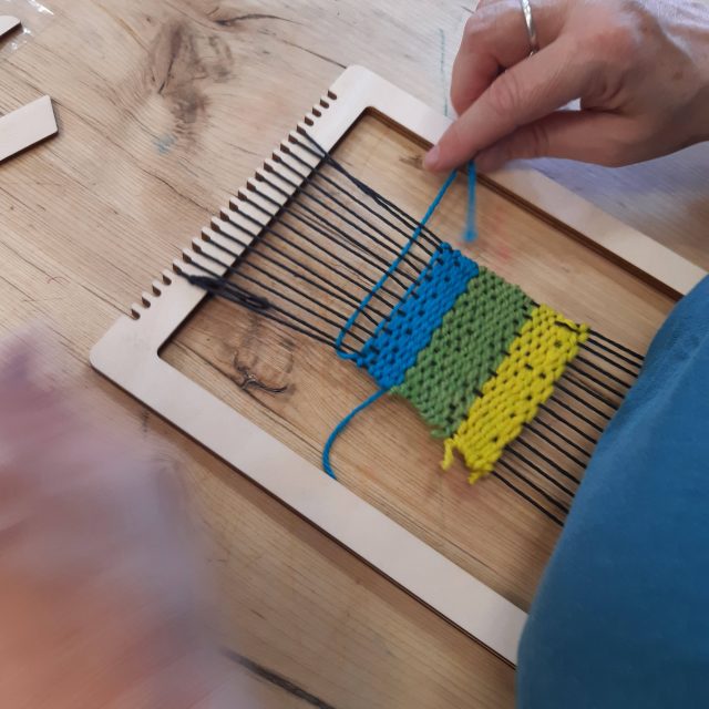 A coloured photograph of a person weaving bright yellow, green and blue yarn.