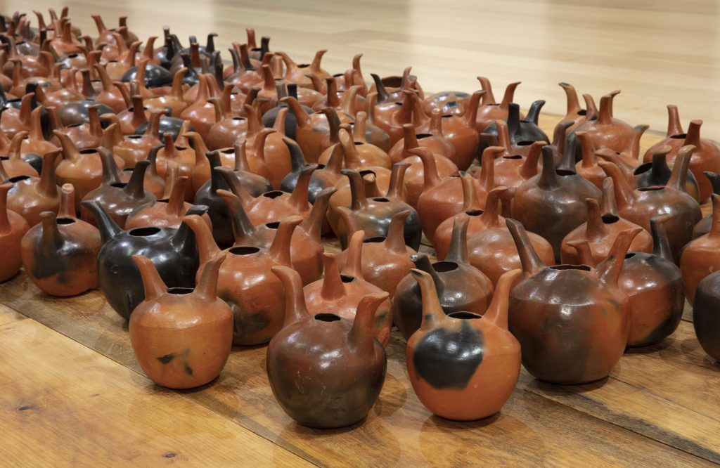 A close up colour photograph of Elias Sime’s ceramic pieces called Bareness, 2014. An installation of multiple ceramic pieces, in shades of orange terracotta clay and some in darker shades. Each piece looks like a pot with two handles with an opening in the middle. 