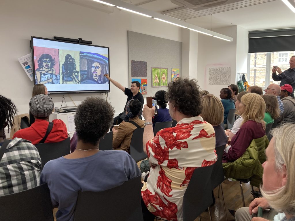 The view is from the back of a room which is packed with many people sitting down, facing the front  and listening to a man that sits at the front of the room, smiling and pointing towards a projector screen showing a series of artworks.