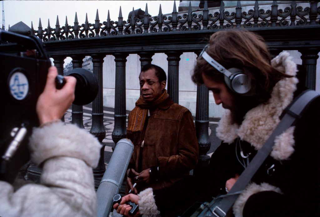 A colour photograph of writer James Baldwin surrounded by press.
