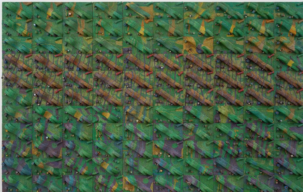 A colour photograph of Elias Sime’s Tightrope: Behind the Processor #6. A three-dimensional wall piece of grids that looks like an ariel view of maps, that has layered texture, made from woven electrical wires and other components, in shades of greens, yellow, blue, purple, brown and red. 
