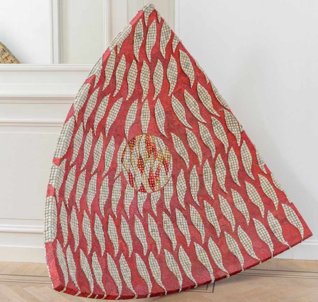 A colour photograph of Elias Sime’s concave floor based sculptural piece called Tightrope: Concave Triangle #2, that is made from beige shade of computer keys in leaf like shapes, and woven reclaimed electrical wires in shades of red, around the computer keys in shades of red. 